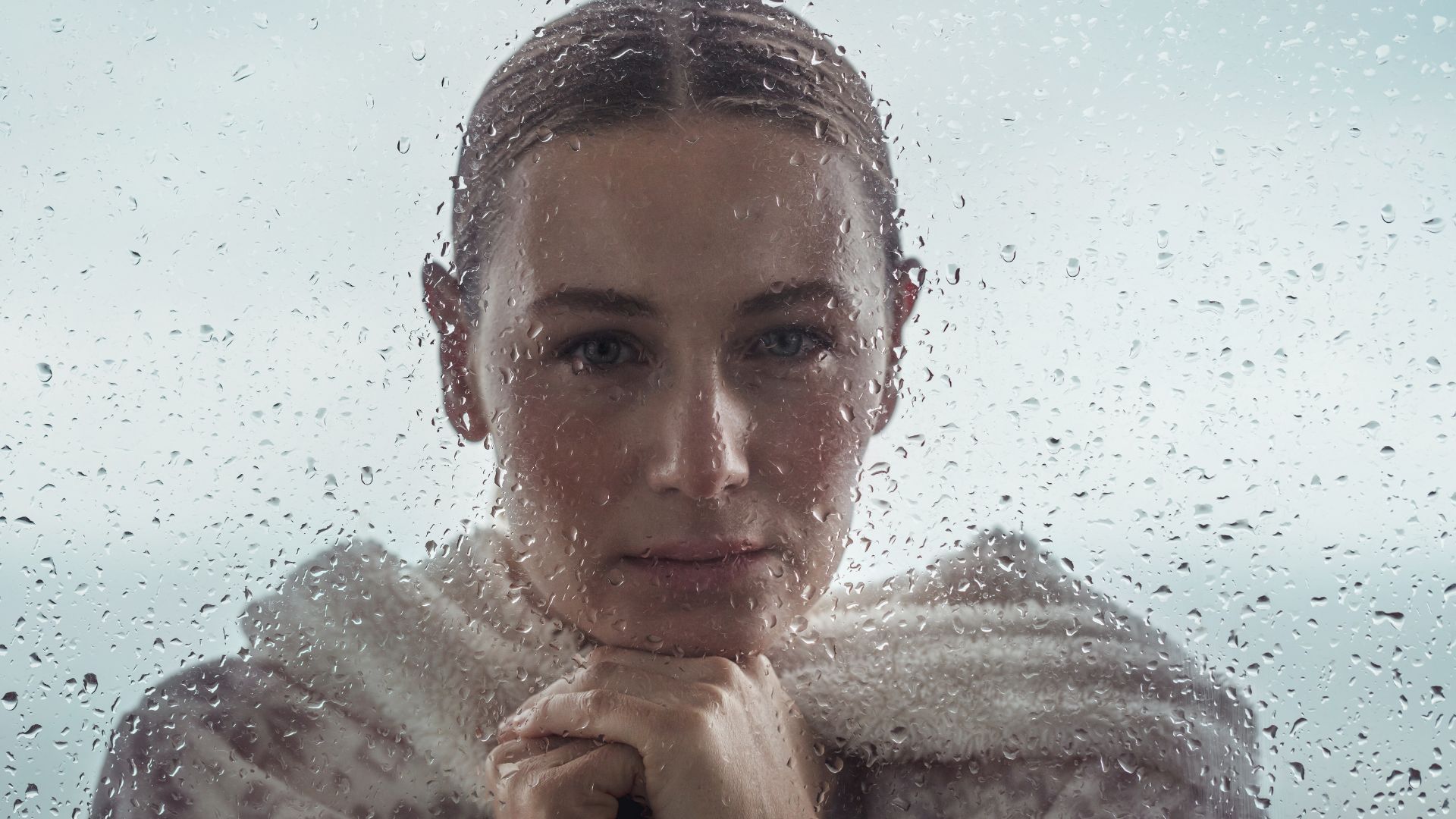 Woman-looking portrait behind a rain-spattered window, hands resting under her chin and a calm, direct gaze.