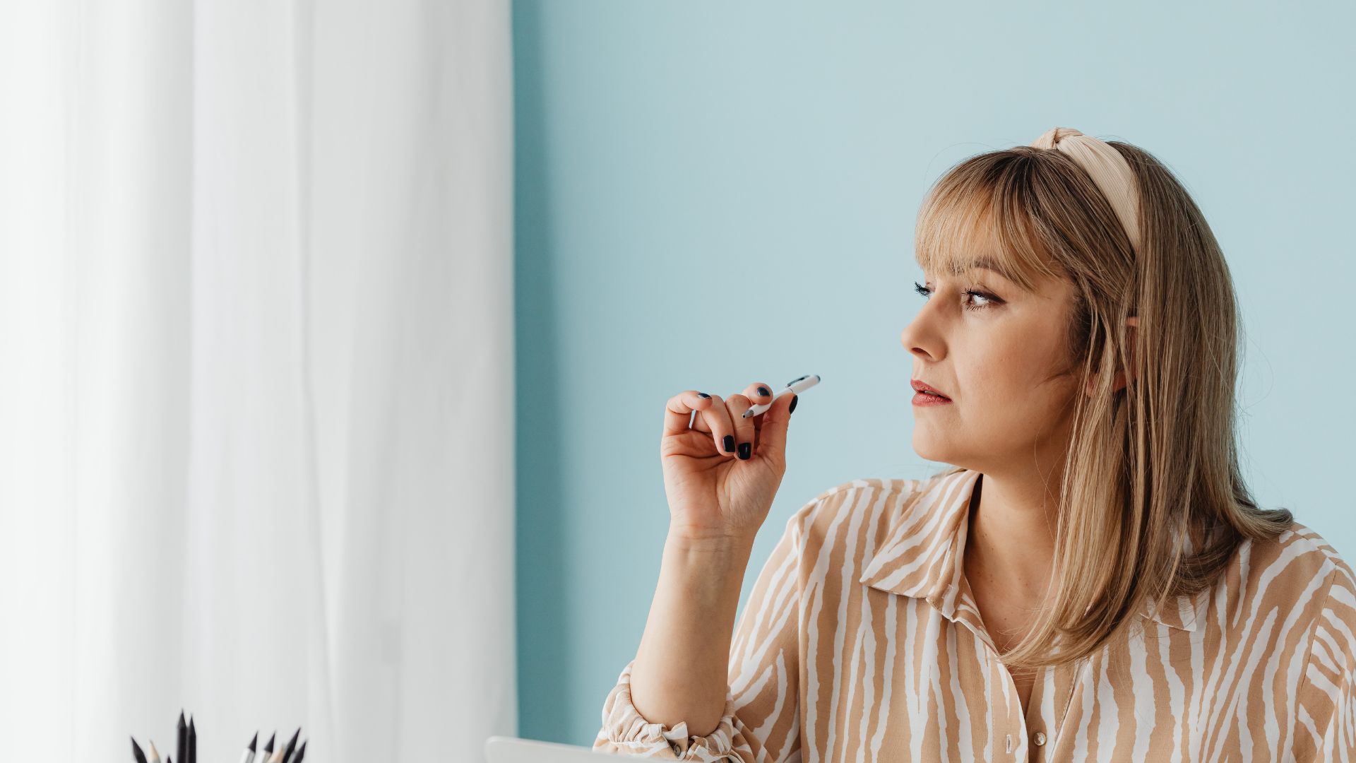 Woman with blonde hair and headband, striped shirt, holding a pen near her lips while looking to the right in a bright blue room with white curtains behind her.