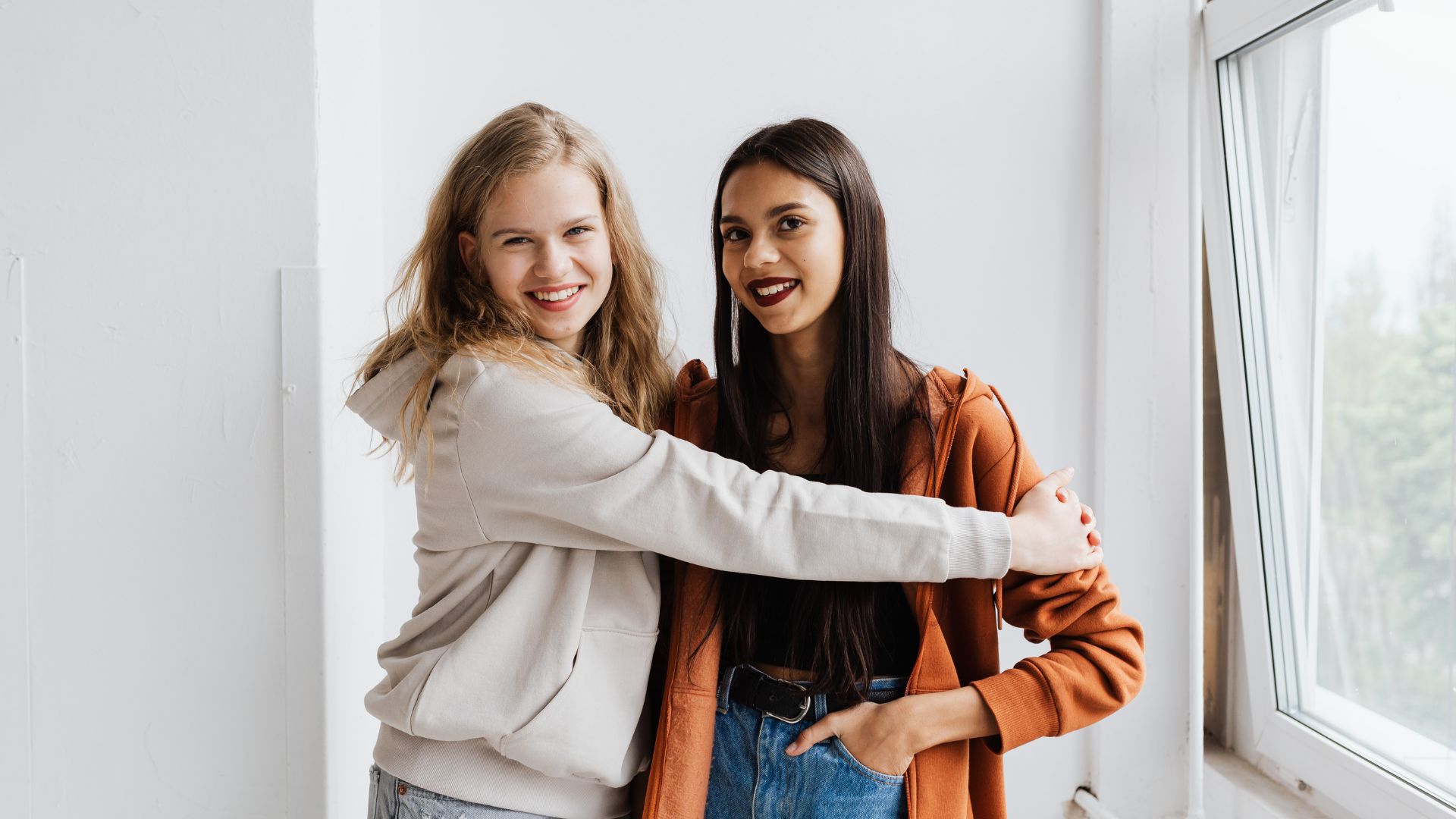 Two young women hugging and smiling by a large window in a bright room, showing warmth and friendship.