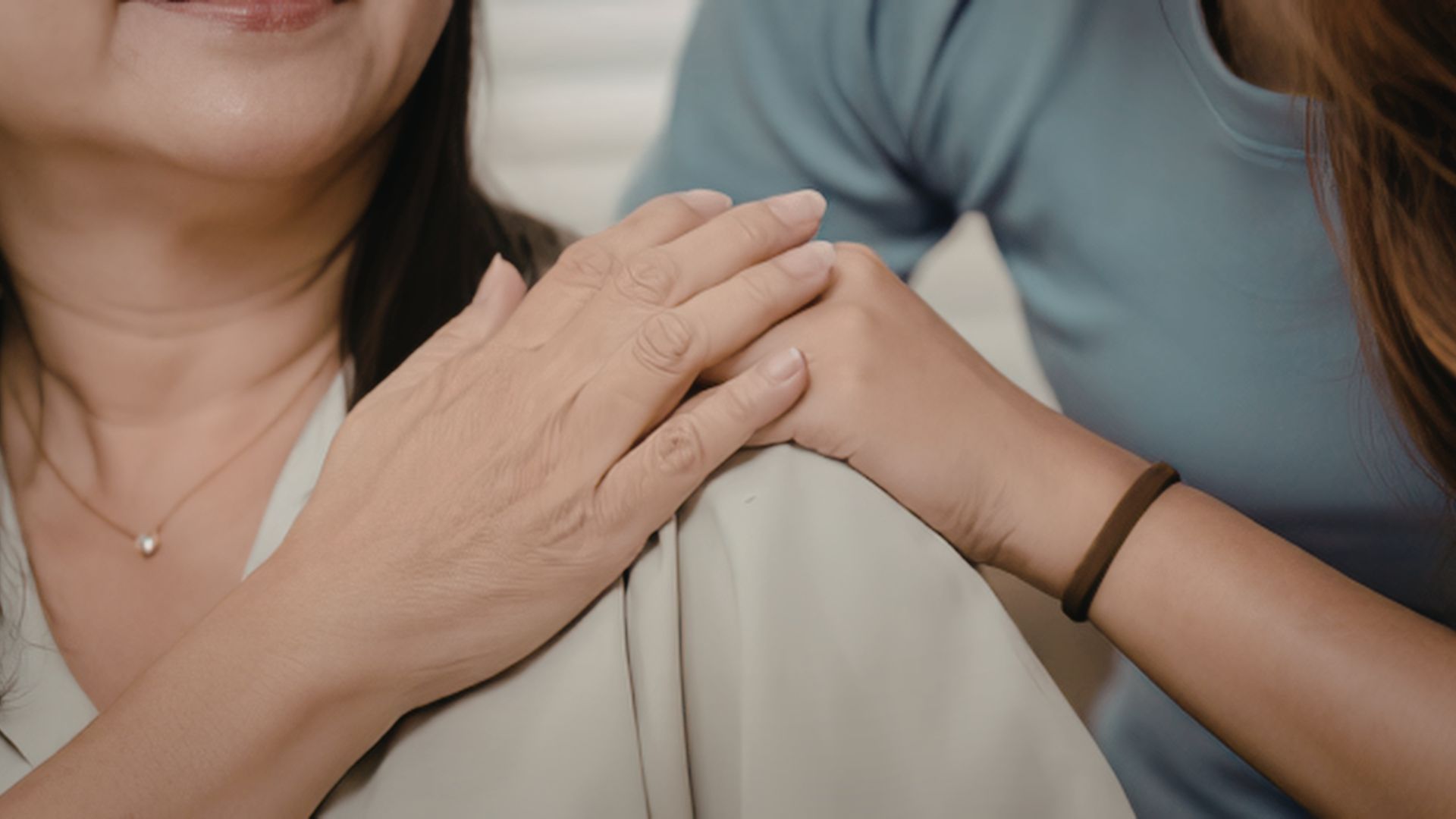 Two people share a comforting touch: a younger person rests their hands on an older person’s shoulder in a supportive hug.